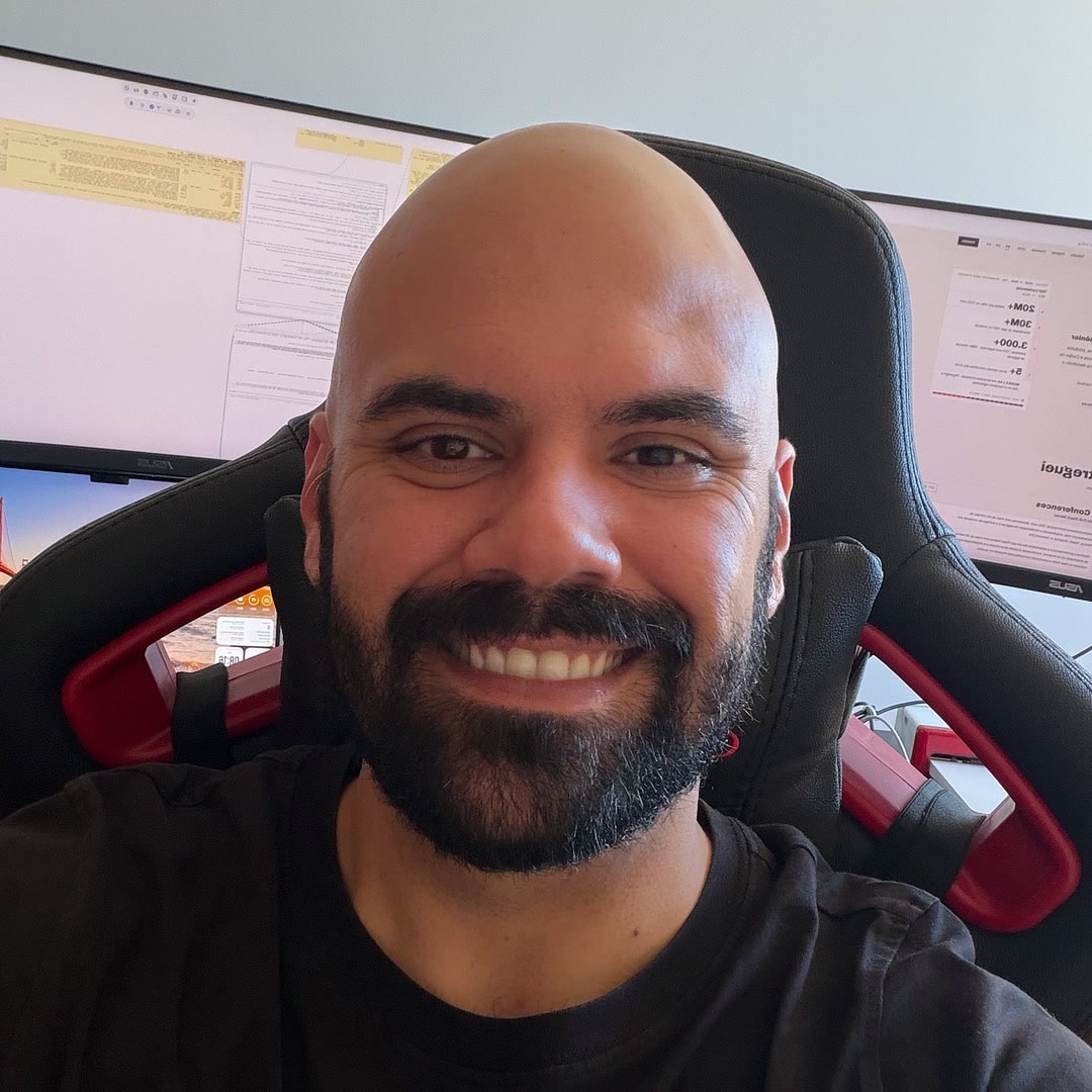 Flavio Gouveia smiling at his desk in a black T-shirt, with his monitors behind him